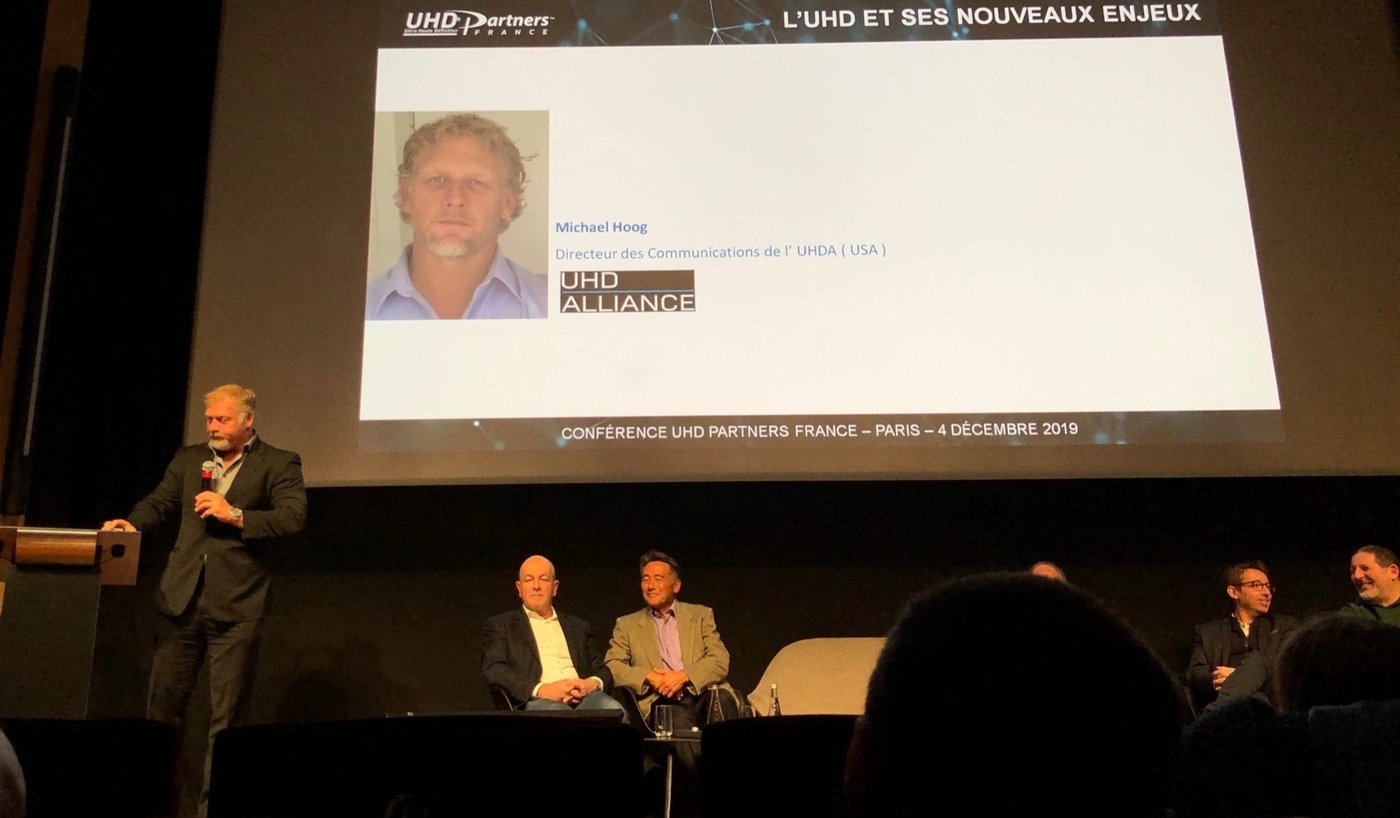 Michael Hoog speaking at a podium at the UHD Partners France conference in Paris, with his name and UHD Alliance title projected on the screen behind him
